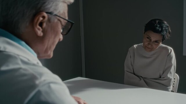 OTS shot of middle aged Caucasian female patient in straitjacket screaming aggressively at senior Caucasian male psychiatrist during medical evaluation. Perfect for mental health awareness campaigns