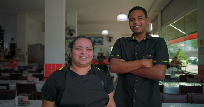 Latin employees smile together inside restaurant dining area, standing in uniform during lighthearted break in casual midday shift at urban food service spot