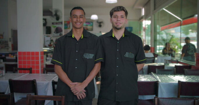 Two restaurant waiters smiling while standing inside dining area, hands folded and behind back, glass wall and tables visible in background