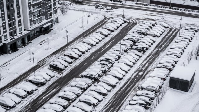 Hundreds of cars are neatly parked in long rows, each one covered in a thick layer of fresh white snow in an outdoor parking area next to a building