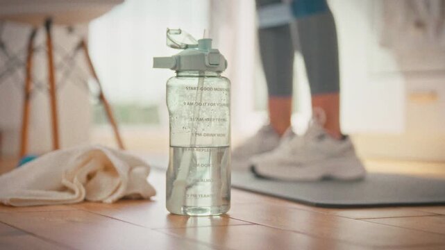In her home kitchen, a young girl stays active, doing squats with elastic bands on her legs for added resistance. Nearby, a sports water bottle keeps her hydrated during her fitness routine