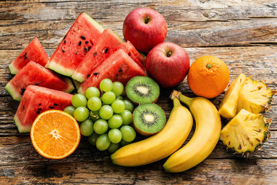 Fresh mixed tropical fruits including watermelon, apples, grapes, kiwi, bananas, orange, and pineapple on wooden surface