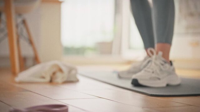 A girl in leggings and white sneakers takes a sports bottle of water against the background of a laid out mat. Concept of proper hydration during home workout during daytime