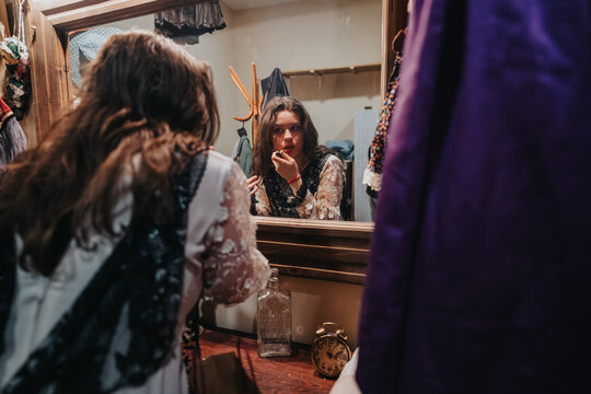 Young actors in a theatre dressing room apply makeup while preparing their costumes. A backstage mirror reflects performers rehearsing and getting ready before a show.