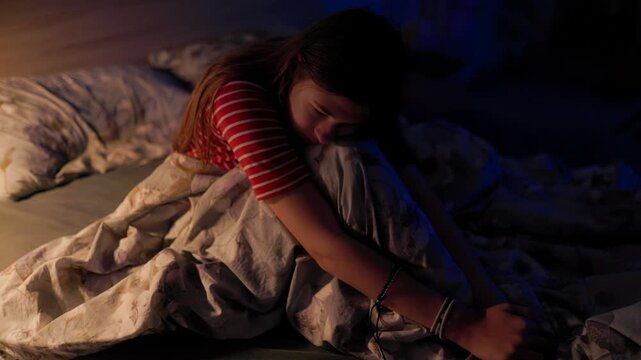 Lonely kid sitting in bed hugging knees feeling sad in dark bedroom interior