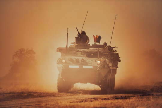 Dramatic shot of an eight-wheeled green military armored vehicle (APC) advancing through a dense dust and haze cloud on a dry dirt field under heavy golden sunset light.