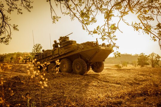 A powerful low-angle shot of a green armored military vehicle (APC) leaping over a dirt ridge, framed by tree branches under a brilliant golden sunset sky.