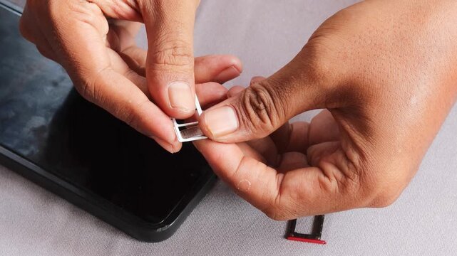 Close-up view of an Asian  man's hands carefully swapping sim card inside smartphone, sitting at table with grey surface with extraction tray nearby during mobile service transition.