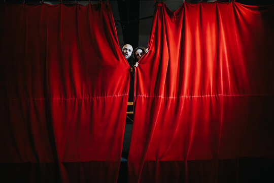 Two young actors peek from behind a red theater curtain. They stand on a dimly lit stage pulling the velvet drape aside.