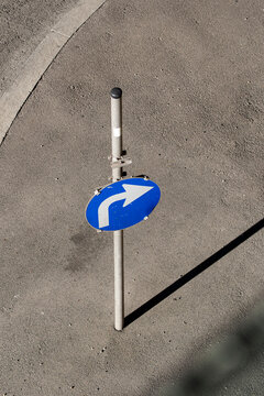 Blue directional sign stands on pavement from above, where arrow symbol, traffic post, shadow and urban road surface create a minimal scene