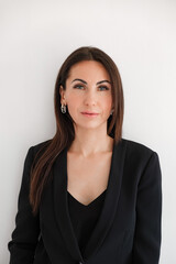 Obraz premium Caucasian woman looking at camera in black formal pantsuit stands against white background. Brunette model with long hair. Business face portrait, formal wear, professional style. Confident person.