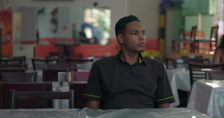 Obraz premium Young restaurant staff member sitting in empty dining area, looking into distance with serene and reflective posture near window