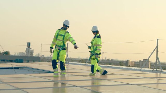 Two engineers in safety clothing with helmet check solar system with city view. Engineers walk alongside rows of solar panels. Man and woman worker inspecting rooftop solar farm at twilight sunset sky