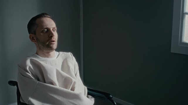 Dolly in shot of Caucasian man wearing straitjacket sitting in wheelchair, staring with shocked expression in psychiatric clinic. Clip is ideal for mental health awareness or medical drama videos