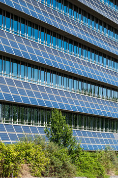 Blue panels run across a building facade with windows and green plants, where urban architecture turns the exterior grid into a patterned surface
