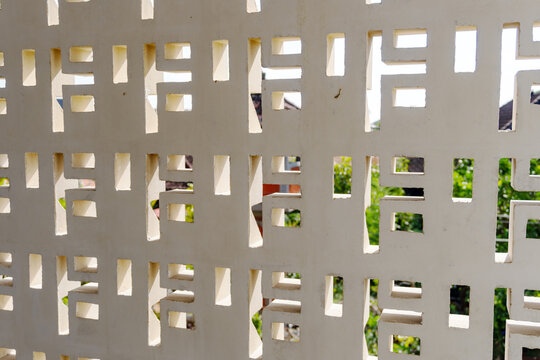 Abstract geometric pattern of a white perforated concrete breeze block wall with natural light and greenery visible through the openings, creating texture, symmetry, and modern architectural detail.