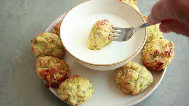 Close-up of a golden crispy baked zucchini tot being dipped into a small bowl of creamy white yogurt sauce