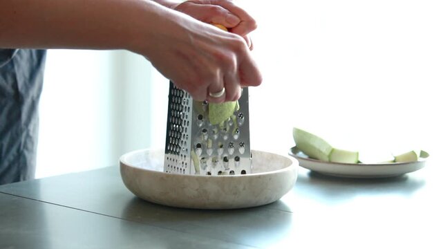Close-up of a woman's hands grating a fresh green zucchini on a metal box grater in a bright kitchen