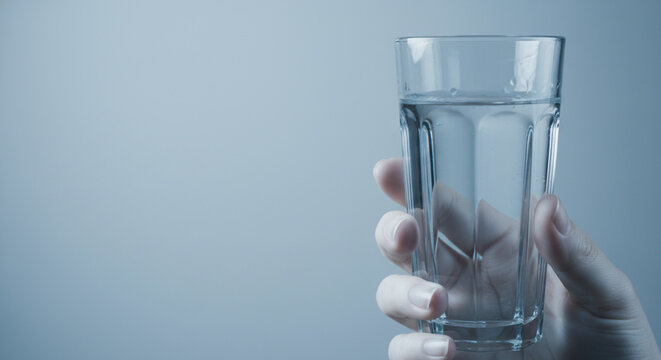 Close up of a hand holding a clear glass filled with clean water against a soft blue background, representing hydration and health