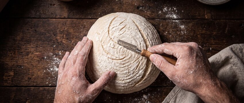 Baker scores sourdough bread dough with lame, preparing loaf for baking, traditional artisan bread making process