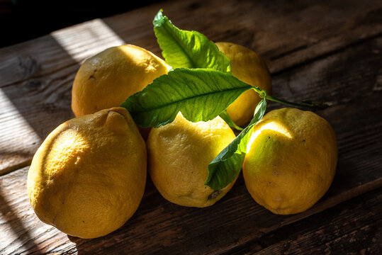 Limones amarillos enteros frescos con hojas verdes sobre mesa de madera r&uacute;stica con luz solar directa y sombras. Toma en plano cenital con iluminaci&oacute;n natural dram&aacute;tica de ventana y sombras marcadas.