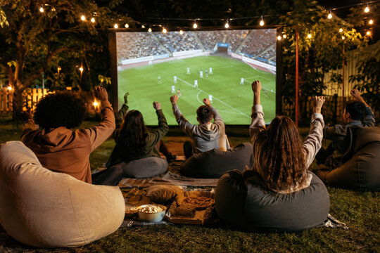 Rear view of friends cheering while watching a soccer match on a large projector screen in a backyard at night. Group of fans celebrating a goal during an outdoor sports watch party.