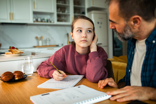 Father helping bored teenage daughter with homework at kitchen table