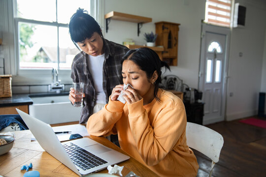 Sick woman working from home in kitchen, partner offering water
