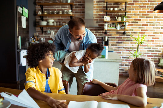 Father lifting daughter while siblings laugh in home kitchen