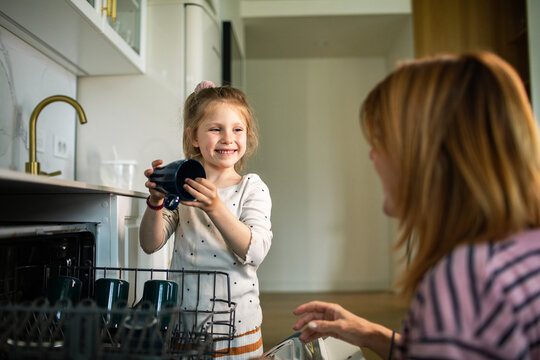 Young girl helping mother load dishwasher in home kitchen