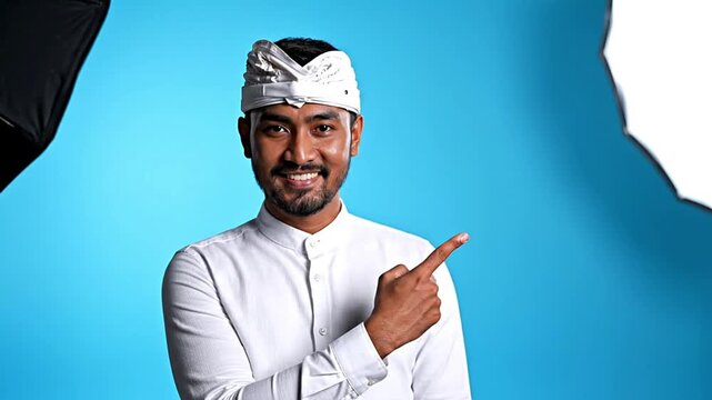 Young Balinese man in traditional Udeng smiles, pointing to space for text or graphics. Ideal for cultural promotions, travel advertisements, or welcoming messages, emphasizing Indonesian heritage...