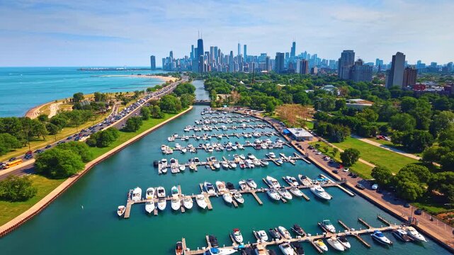 Flight over the vessels at the berths on the blue waterscape. A highway with hectic traffic passes along the yacht club. Chicago downtown skyline in the backdrop.