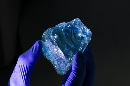 A piece of transparent glass crystal held by hand in blue rubber gloves at a laboratory ready for a test, on dark background