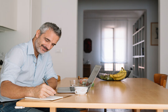 Smiling man writing in notebook and using laptop in home office