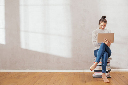 Young woman sitting on stack of books using laptop at home