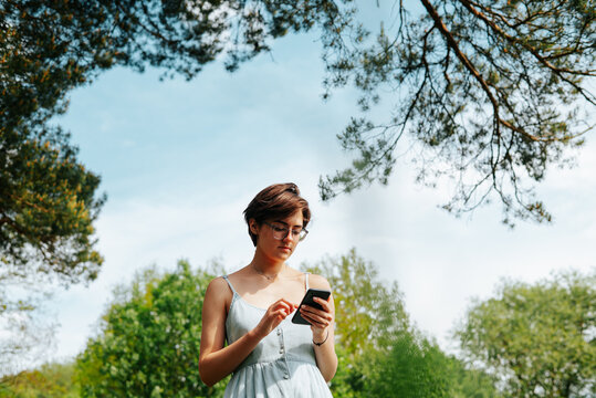 A young woman stands in a park looking at her smartphone while surrounded by trees and green plants. She appears focused and engaged with her device in this natural setting