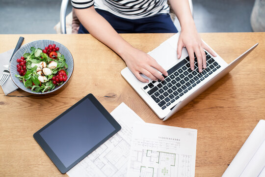 Woman at desk using laptop next to construction plan and salad