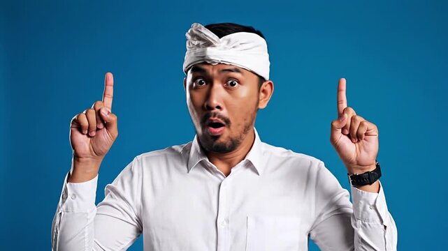 A surprised Asian man in Balinese traditional headwear, 'udeng', points excitedly upwards, highlighting a new idea, amazing discovery, or unexpected solution.