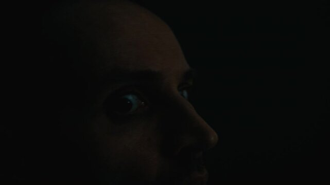 Side view closeup of adult Caucasian man experiencing panic attack and staring with wide terrified eyes in dark room. Footage is ideal for mental health awareness and PTSD educational videos
