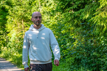 Naklejka premium Cheerful portrait young bald man glasses white hoodie, park alley trees green background, smiling winking face portrait happy expression, casual style and active lifestyle summer day outdoor sunlight
