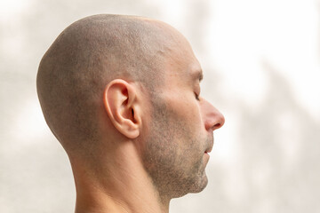 Fototapeta premium Side profile of a bald man with closed eyes, calm expression, shaved head, stubble, white background, meditation, relaxation, peace, contemplation, portrait close up