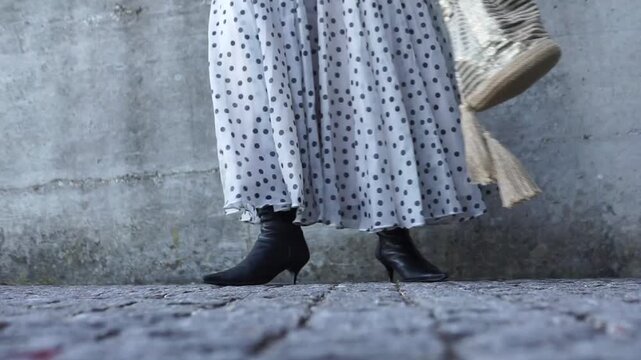 A low-angle side tracking shot showing a woman's legs in black high-heel boots and a polka dot skirt walking past the camera along a concrete wall.
