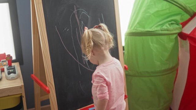 Little blonde girl in pink shirt erases chalkboard with hand at wooden easel inside building playroom with tent and storage; childhood concentration.