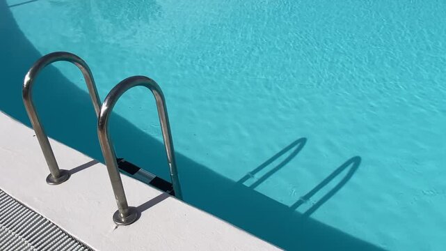 Cinematic video of a stainless steel swimming pool ladder beside crystal-clear blue wate. Relaxing summer travel and leisure concept ideal for luxury resort, vacation, wellness and hospitality themes
