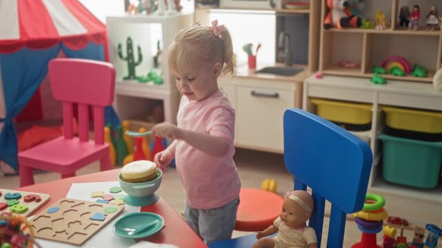 Little blonde girl feeds baby doll with toy spoon at a small table in a colorful playroom; gentle nurturing play.