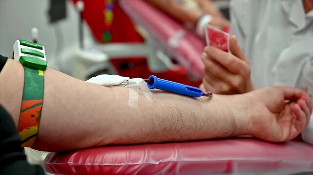 Close-up of a secured needle taped in a blood donor arm with a blue clamp while a nurse's hands in the background handle a sample tube and hold a special medical blood collection bag at a clinic