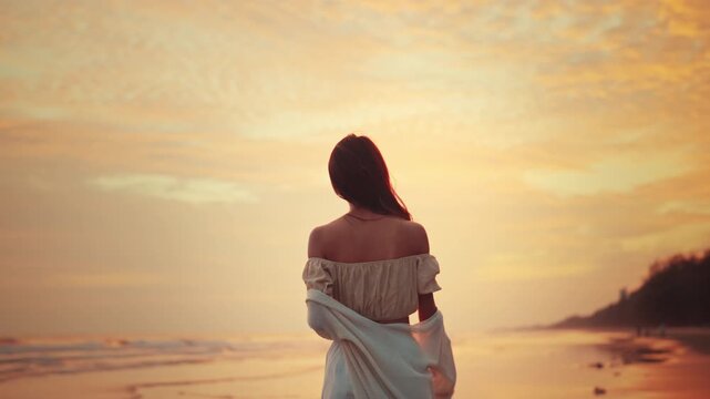 Rear view of happy young Asian woman walking on the beach, looking away at the golden sunset sky. Beautiful female traveler enjoying summer vacation holiday, feeling freedom and carefree mindfulness