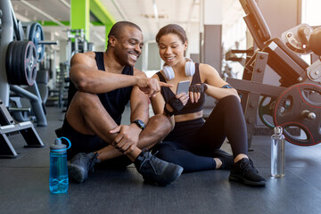 Naklejka premium Young black sitting on the gym floor reviewing workout information on a smartphone. Fitness tracking, recovery, teamwork, and healthy lifestyle concept