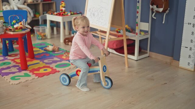 Little blonde girl rides wooden tricycle in building playroom amid colorful foam number mat and scattered toys; childhood joy.