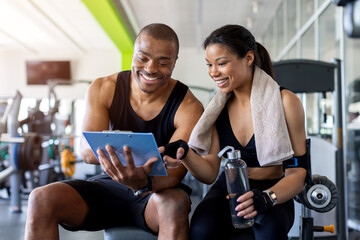 Smiling trainer and client reviewing workout information together in modern fitness center,...